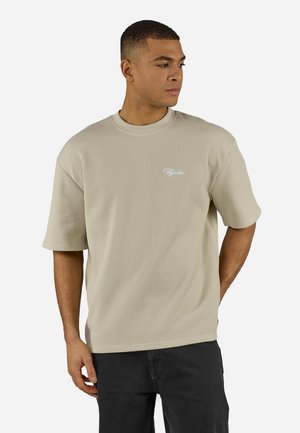 Mies, joka käyttää beigeä, ribbineulosta lyhythihaista paitaa, jossa on pieni valkoinen kirjallinen logo rinnassa, seisoo vasen käsi selän takana, katsoo oikealle.