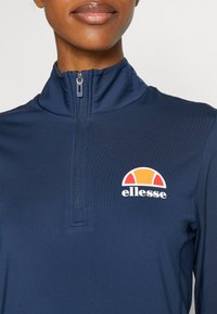 Mörkblå, zip-up tröja med hög krage, utrustad med en kontrasterande färgglad logotyp på vänster bröst. Slät, strukturerad tyg.