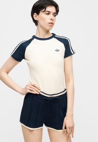 Γυναίκα που φοράει μπλουζάκι Adidas σε κρεμ και ναυτικό μπλε με ταιριαστό σκούρο μπλε πλεκτό σορτς, στέκεται με το ένα χέρι στη μέση της μπροστά σε απλό φόντο.
