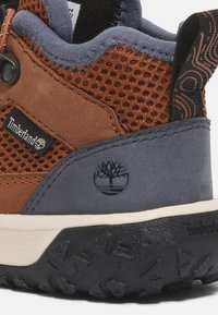 Zapatilla marrón y gris con parte superior de malla, cuello acolchado, suela de goma, acentos texturizados y parche de logo. Presenta materiales duraderos para uso al aire libre.