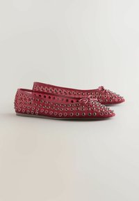 Une paire de ballerines rouges décorées de œillets argentés et de petits nœuds à l'avant, présentées côte à côte sur un fond uni.