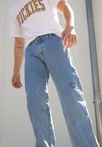 Lys blå denim cargo bukser med vide ben, med knælapper og et lille logo. Kombinert med en hvid T-shirt med "DICKIES" i gult.