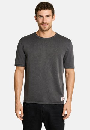 Mann mit kurzen lockigen Haaren, der ein schlichtes dunkelgraues kurzärmliges T-Shirt und schwarze Hose trägt und in die Kamera lächelt.