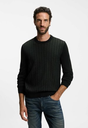 Uomo con capelli scuri ricci e barba, che indossa un maglione nero con fantasia e jeans blu, in piedi con una mano in tasca su sfondo bianco.