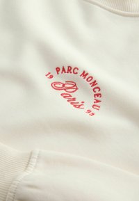 Valkoinen collegepaita, jossa on punainen brodeerattu kuvio, jossa lukee "PARC MONCEAU Paris 19 95". Hihansuissa on ribbireunus ja pinta on sileä.