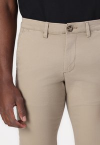 Pantalones de algodón beige con corte recto, que cuentan con cremallera y botón en la parte frontal, bolsillos laterales y costuras visibles para mayor durabilidad.