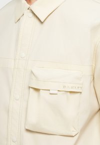 Ανοιχτό μπεζ μπουφάν φτιαγμένο από λείο ύφασμα, με κουμπωτή σχεδίαση, γιακά και μπροστινή τσέπη με ανάγλυφο λογότυπο της μάρκας.