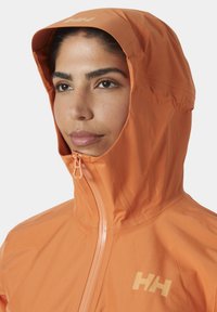Chaqueta ligera con capucha en color naranja y cremallera frontal, que presenta un logo sutil y una textura suave, diseñada para uso al aire libre.