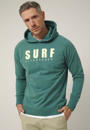 Hombre con cabello castaño claro que lleva una sudadera con capucha color azul verdoso con el texto "SURF ALTONADOCK", pantalones beige, de pie con las manos en los bolsillos contra un fondo sencillo.