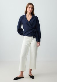 Camicia avvolgente blu navy con un nodo in vita, maniche lunghe e colletto, abbinata a pantaloni bianchi a vita alta e a gamba larga e ballerine nere.