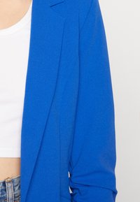 Blå kavaj med en slät textur, skräddarsydd design och en enkel knappstängning, kombinerad med en vit kortärmad topp.