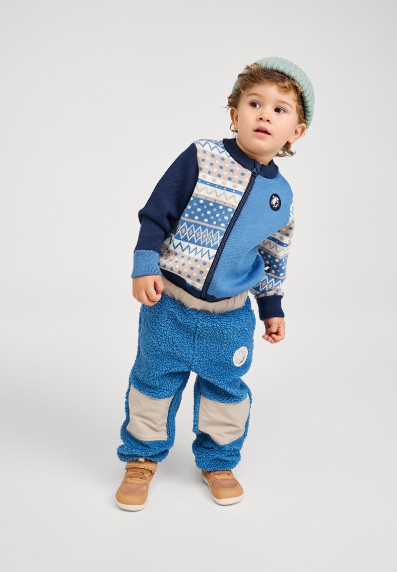 Tout-petit portant un pull à fermeture éclair bleu à motifs, un pantalon en polaire bleu avec des patchs beige, des chaussures couleur cuir clair et un bonnet tricoté vert clair, debout sur un fond uni.