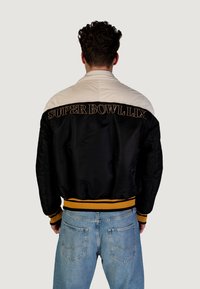 Giacca bomber nera in satin con spalle color crema, "SUPER BOWL LIX" ricamato sul retro, polsini e orlo a coste gialli, texture liscia.