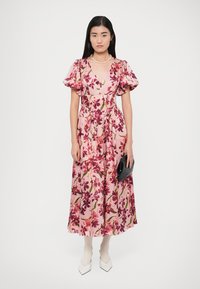 Floral maxi dress in pink with puff sleeves, featuring a tied waist and vivid burgundy flowers. Model holds a black clutch, wears white shoes.