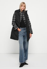 Svart quiltet puffervest med hette, brukt over en svart topp og grå rutete skjorte, kombinert med vide blå jeans og svarte sko.