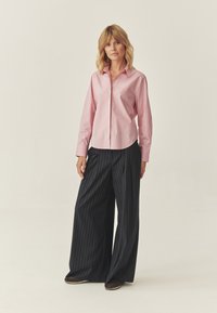 Jasnoróżowa koszula z guzikami i kołnierzykiem, zestawiona z wysokimi, szerokimi spodniami w ciemnoniebieskie paski. Całość dopełniają brązowe buty.