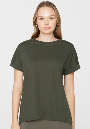 Joven con cabello rubio liso, vistiendo una camiseta de manga corta de color verde oliva lisa, de pie frente a un fondo blanco.