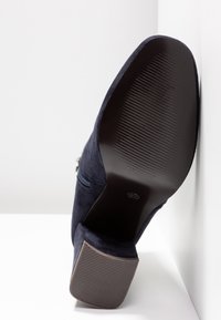 Navy blue suede high heel shoe with a textured rubber sole, featuring a silver zipper detail on the side and a rounded toe design.