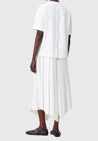 Mannequin portant un top blanc à manches courtes avec une fermeture éclair dans le dos et une jupe midi plissée blanche avec un ourlet asymétrique, ainsi que des ballerines noires cloutées.