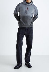 Felpa grigia con tasca frontale e logo rosso, abbinata a jeans scuri e stivali in pelle nera, con una vestibilità rilassata e una texture liscia.