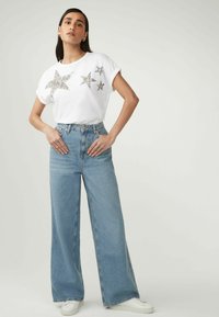 T-shirt en coton blanc avec des sequins en forme d'étoile argentés sur le devant, associé à un jean en denim large taille haute bleu clair et des baskets blanches.