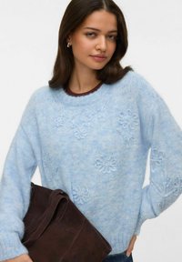 Gaiši zilā adītā džemperī ar ziedu izšuvumu, apaļu kakla izgriezumu, brīvu piegriezumu un ribotām aprocēm. Modeles rokās ir tumši brūna zamšādas soma.