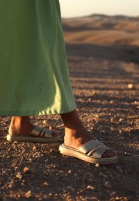 Světlé béžové skluzné sandály s hladkým povrchem, které mají dva křížící se popruhy. Nosené na holé noze, umístěné na texturovaném terénu.