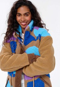 Kobieta z długimi kręconymi włosami uśmiechająca się, ubrana w beżową polarową kurtkę w niebieskie i fioletowe wzory, z rękami skrzyżowanymi.