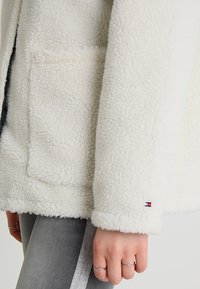 Witte fleece jas met een gestructureerd, pluche oppervlak, voorzien van een voorkantzak en een klein logo op de manchette. Zacht, knus ontwerp.