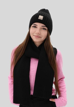 Beanie nero a maglia con un'etichetta e sciarpa nera abbinata. Il modello indossa un maglione rosa pallido con maniche lunghe. Texture semplice e a coste.