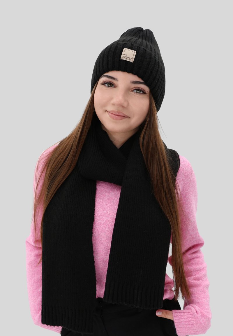 Zwarte gebreide beanie met een label en bijpassende zwarte sjaal. Het model draagt een lichtroze trui met lange mouwen. Eenvoudige, ribbelstructuur.