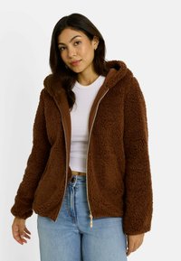 Veste à capuche zippée marron en tissu moelleux et texturé, avec un intérieur contrastant de couleur marron. Portée sur une chemise blanche et un jean en denim.