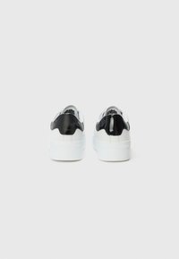 Baskets blanches avec des accents en cuir verni noir, une texture lisse, une forme arrondie et de grosses semelles blanches. Logo de la marque embossé sur le côté.