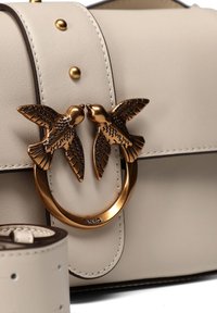 Μπεζ δερμάτινη τσάντα χειρός με κυκλικό μπρούτζινο κούμπωμα που απεικονίζει δύο πουλιά που φιλιούνται. Περιλαμβάνει ταιριαστό μπεζ λουρί με διακοσμητικές τρύπες.