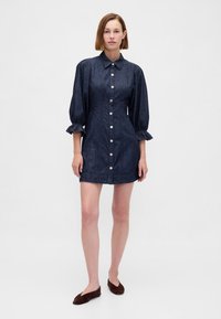 Rochie din denim albastru închis, cu guler, mâneci lungi bufante și închidere cu nasturi în față. Prezintă manșete adunate și o siluetă cambrată.