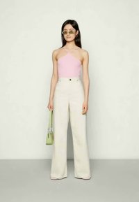 Top halter rosa claro com alças cruzadas, emparelhado com calças largas brancas de cintura alta. A modelo segura uma pequena bolsa verde.