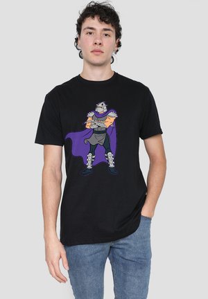 Young man wearing a black t-shirt with a cartoon armored warrior in a purple cape standing with crossed arms on the front.