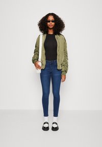Casaco bomber verde-oliva com mangas franzidas, body preto, jeans skinny azuis, meias brancas, sapatos de plataforma pretos e uma pequena mala.