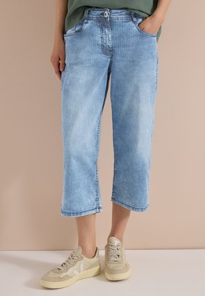 Persona che indossa jeans corti a righe verticali azzurro chiaro e sneakers beige, in piedi davanti a uno sfondo neutro.