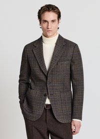 Blazer a scacchi marrone e blu realizzato in tessuto testurizzato, con chiusura a singolo bottone e due tasche laterali, indossato sopra un dolcevita crema.