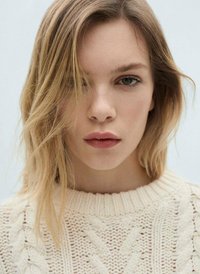 Maglione intrecciato di colore beige chiaro con un ampio scollo a girocollo. Il modello ha capelli biondi ondulati e lunghi fino alle spalle e un trucco naturale.