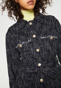 Femme portant un manteau bleu marine texturé avec des boutons dorés, des poches avant avec détail frangé, une ceinture nouée à la taille, et un col roulé vert clair en dessous.