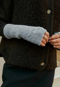 Graue gerippte Strick-Armwärmer mit dekorativem Schlaufen-Detail, kombiniert mit einem strukturierten schwarzen Überbekleidungsstück mit großen Knöpfen.