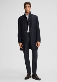 Marineblauwe wollen overjas met hoge kraag, voorzien van knoopsluiting en voorzakken, gecombineerd met donkere geruite broeken en bruine leren schoenen.