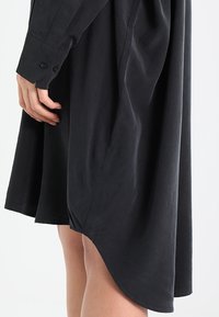 Schwarzes Kleid mit lockerem Schnitt und geschwungenem Saum. Hergestellt aus leichtem Stoff. Mit langen Ärmeln und Knopfdetails sowie einer entspannten Silhouette.