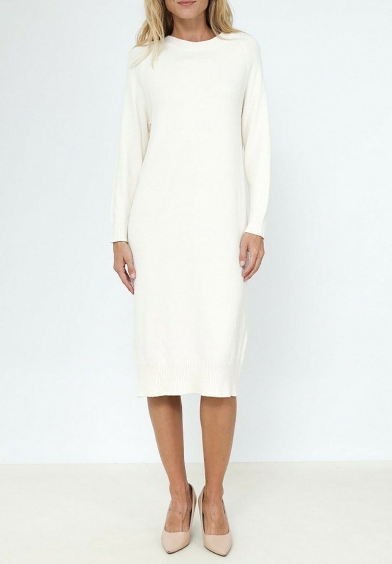 Robe en tricot crème à manches longues, col rond, avec des poignets et un ourlet côtelés. Portée avec des chaussures à talons beiges. Texture lisse, design mi-long.