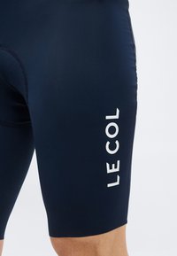 Detailný záber na tmavomodré cyklistické šortky na stehne osoby s bielym vertikálnym nápisom "LE COL" na boku.