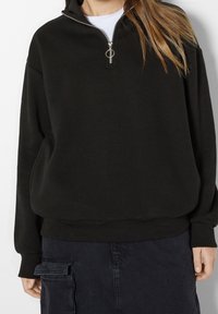 Svart sweatshirt med hög krage och halv dragkedja, med en avslappnad passform och ribbade ärmslut. Slät textur och minimalistisk design.