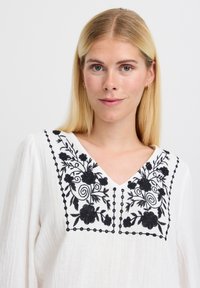 Blusa bianca con ricami floreali neri attorno a un neckline a V e tessuto texturizzato, con accenti decorativi di perline nere sui ricami.