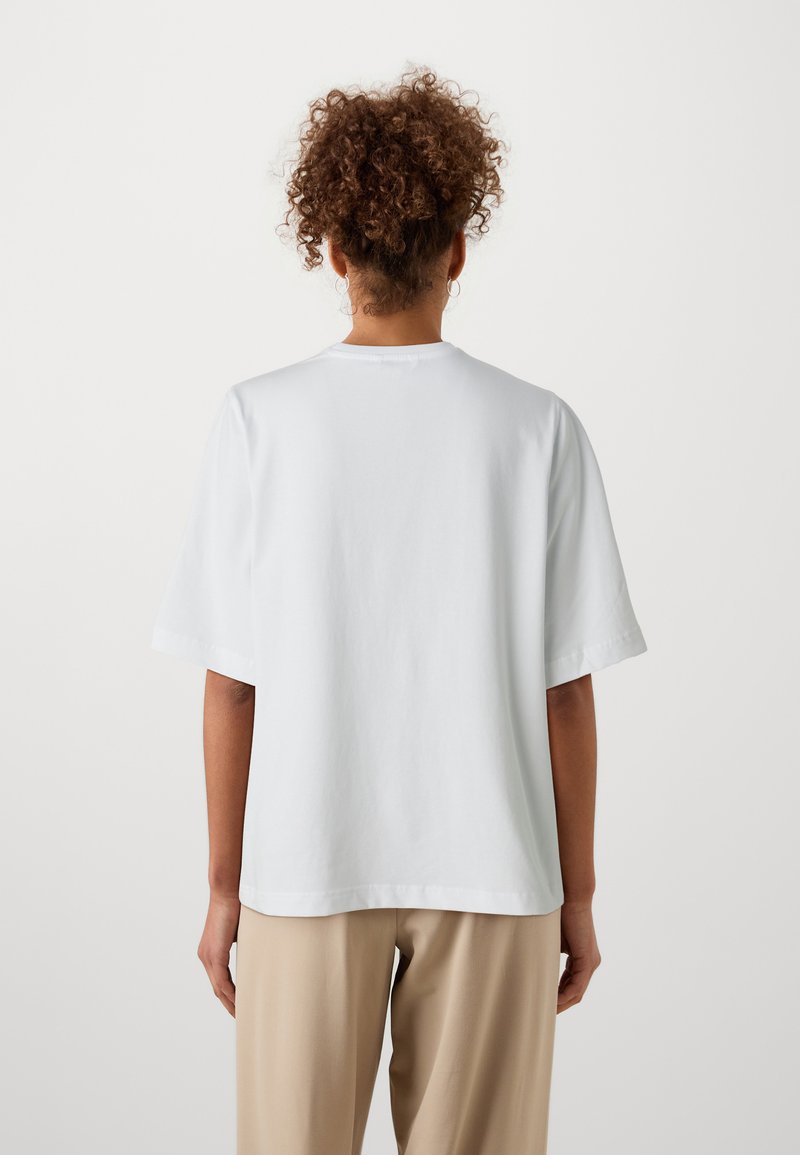 Vit bomull t-shirt med korta, breda ärmar och rund halsringning. Bakifrån visar den en jämn textur och en avkopplad passform, tillsammans med beiga byxor.